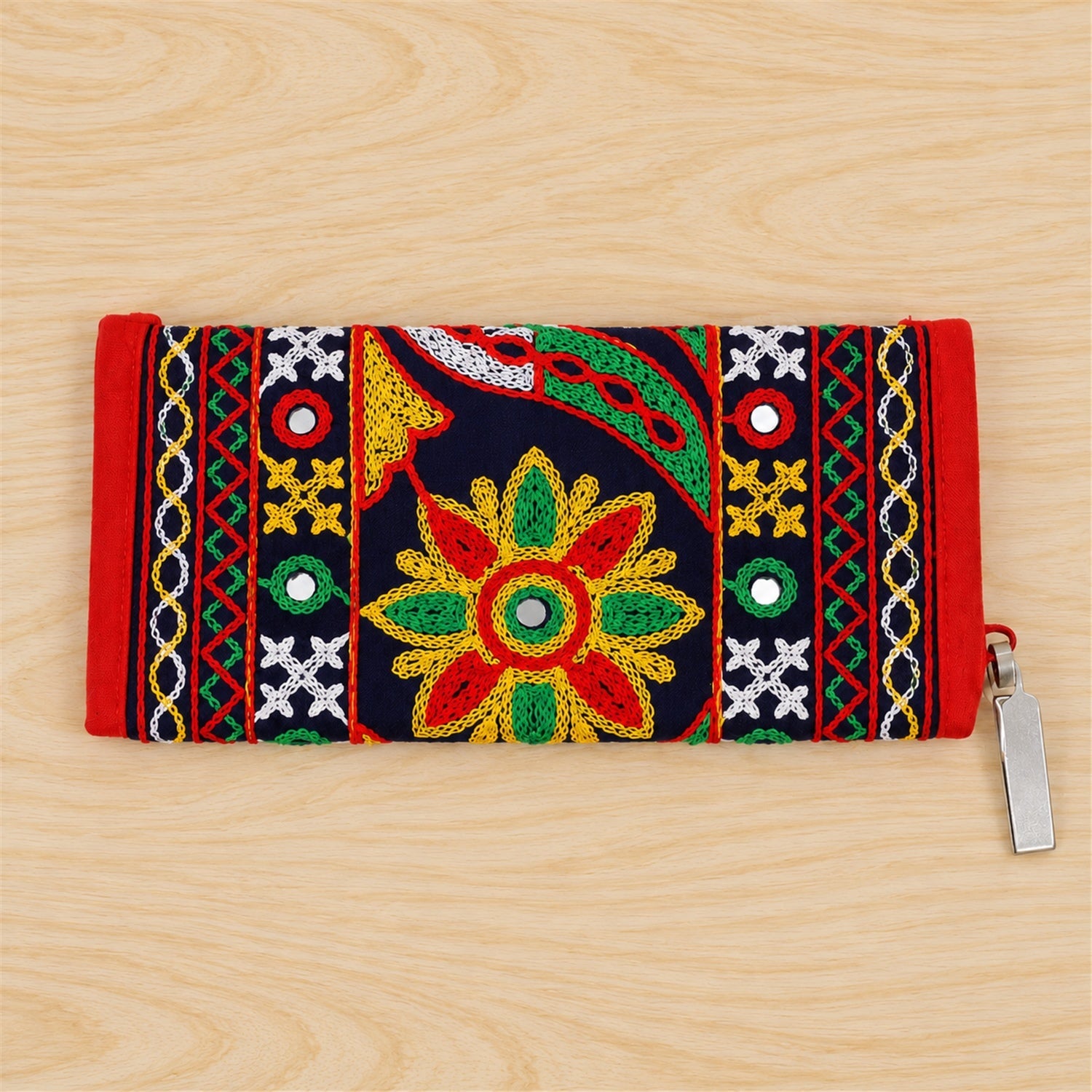 The embroidered clutch, featuring a red and dark blue design with colorful stitching and small mirrors, lies flat on a light