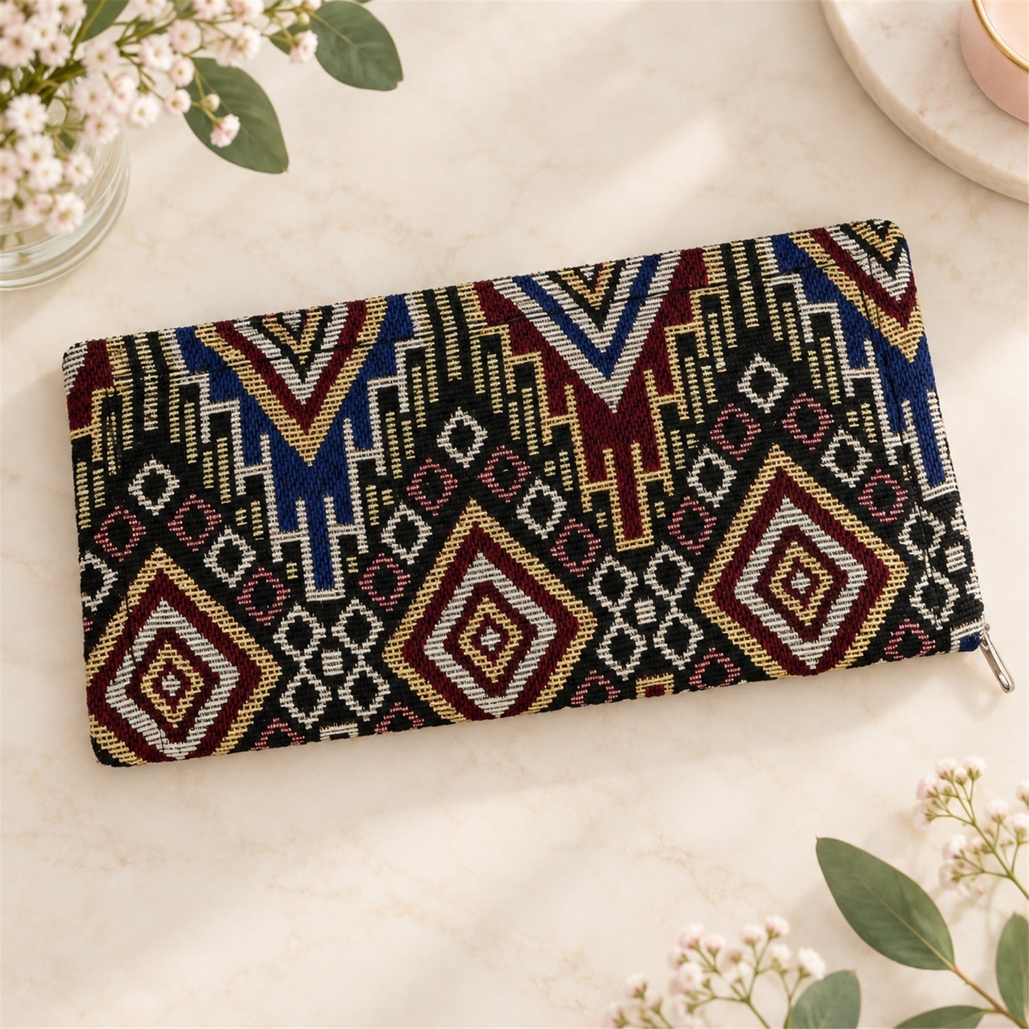 A closed geometric patterned clutch wallet lies on a white marble surface, surrounded by delicate white flowers and green