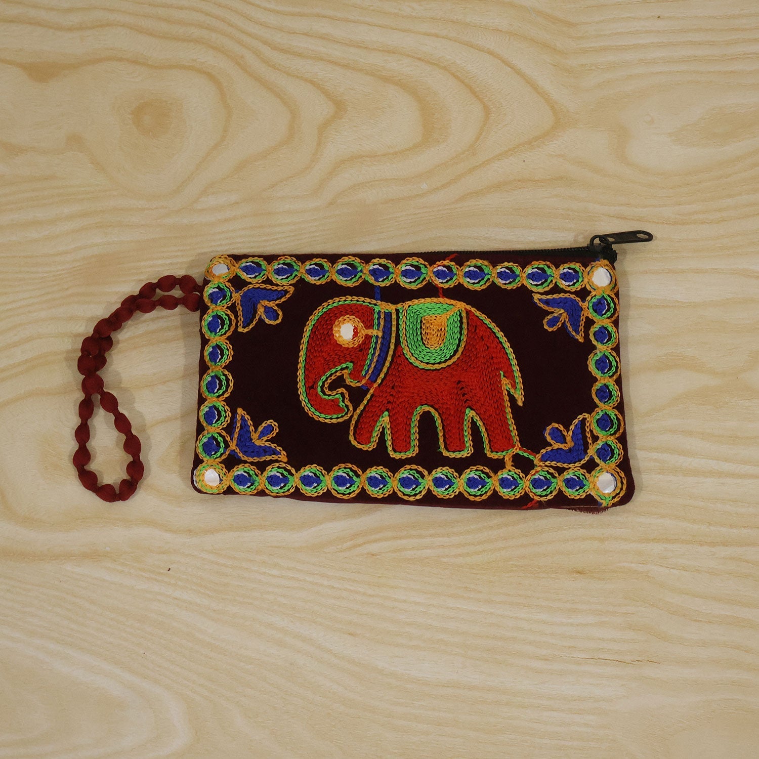 A dark red velvet clutch purse with an embroidered red elephant and blue and green beaded trim, resting on a light wooden