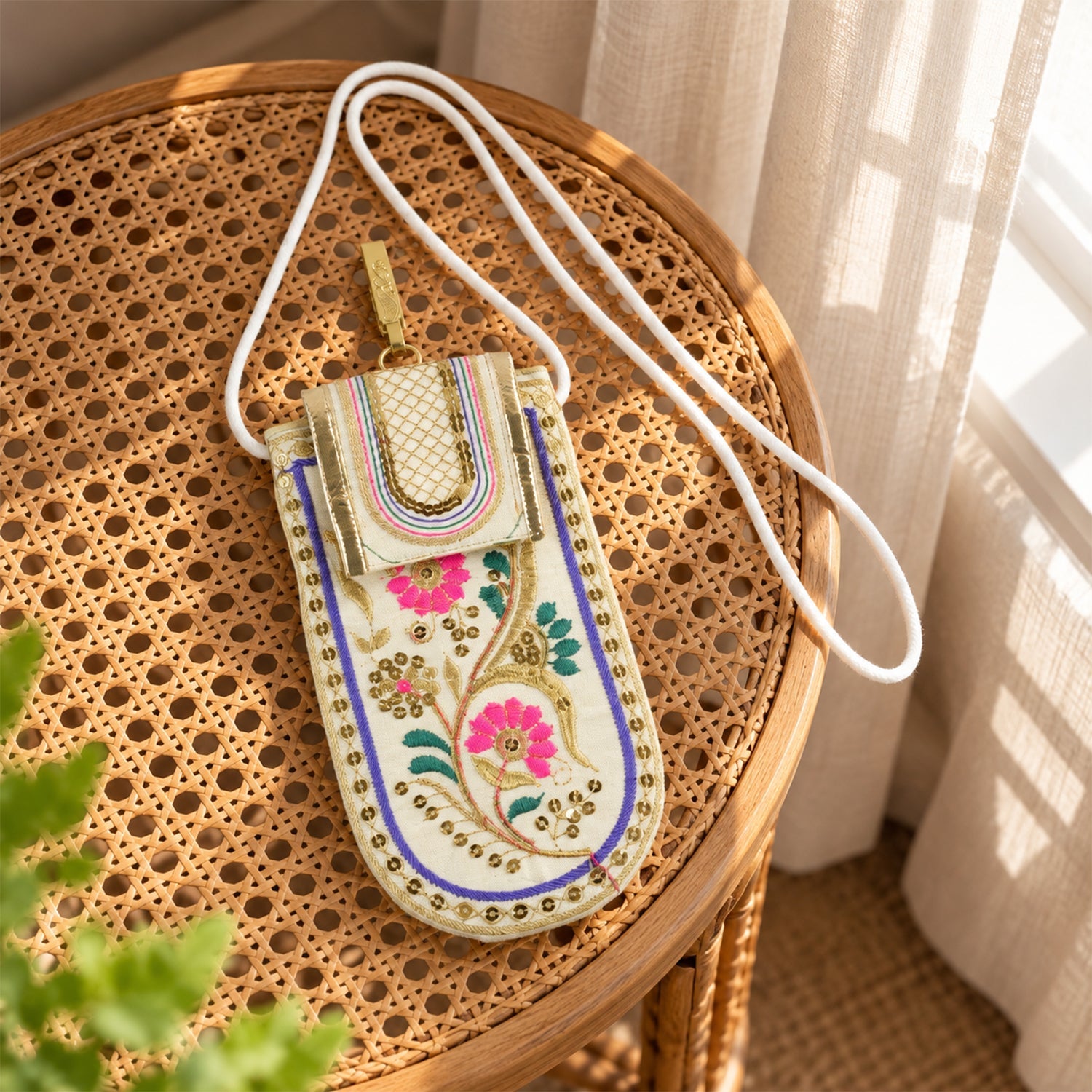 An embroidered mobile pouch with a white sling strap rests on a woven rattan table near a window.