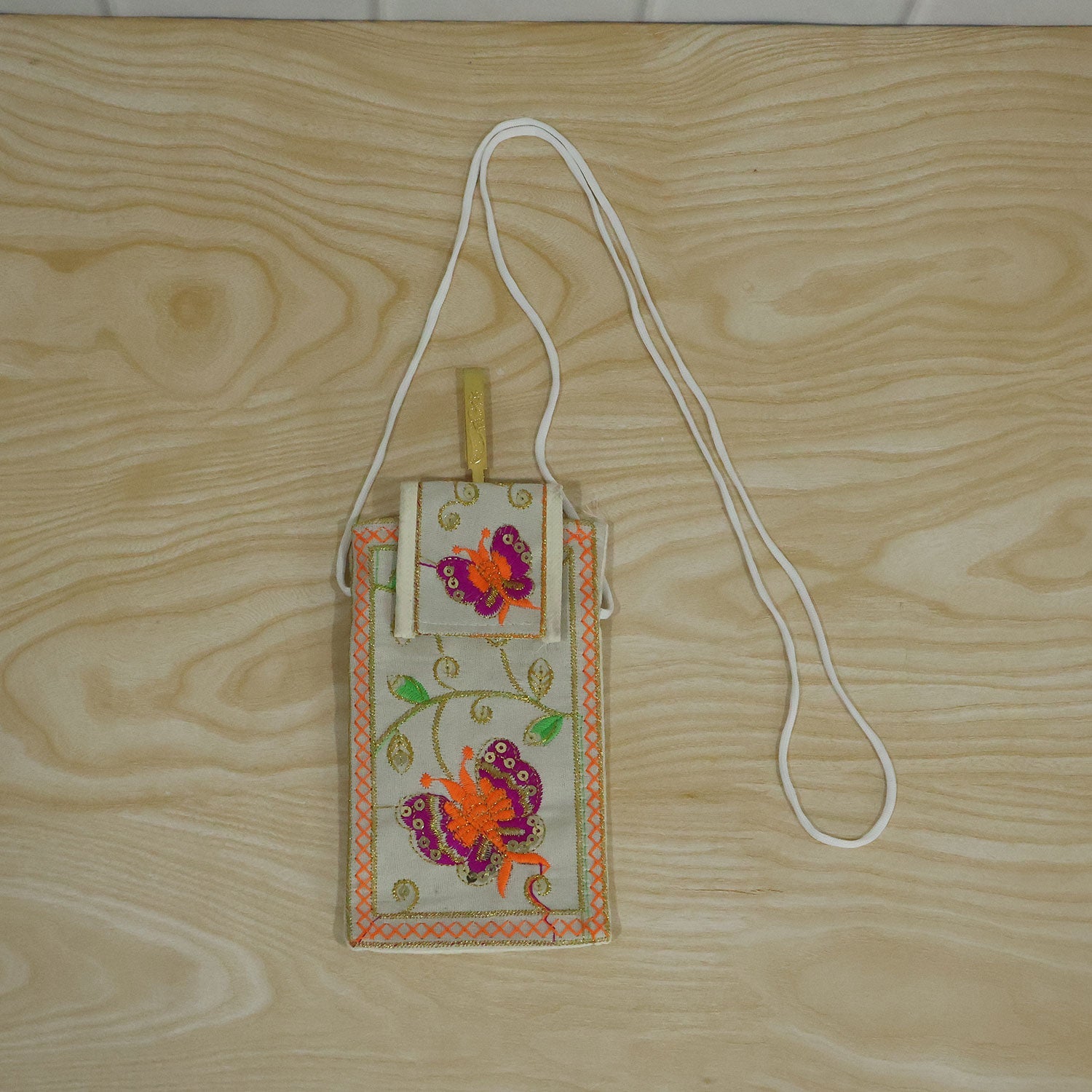 White embroidered mobile pouch with orange and pink butterfly design and white sling strap, lying on a wooden surface.