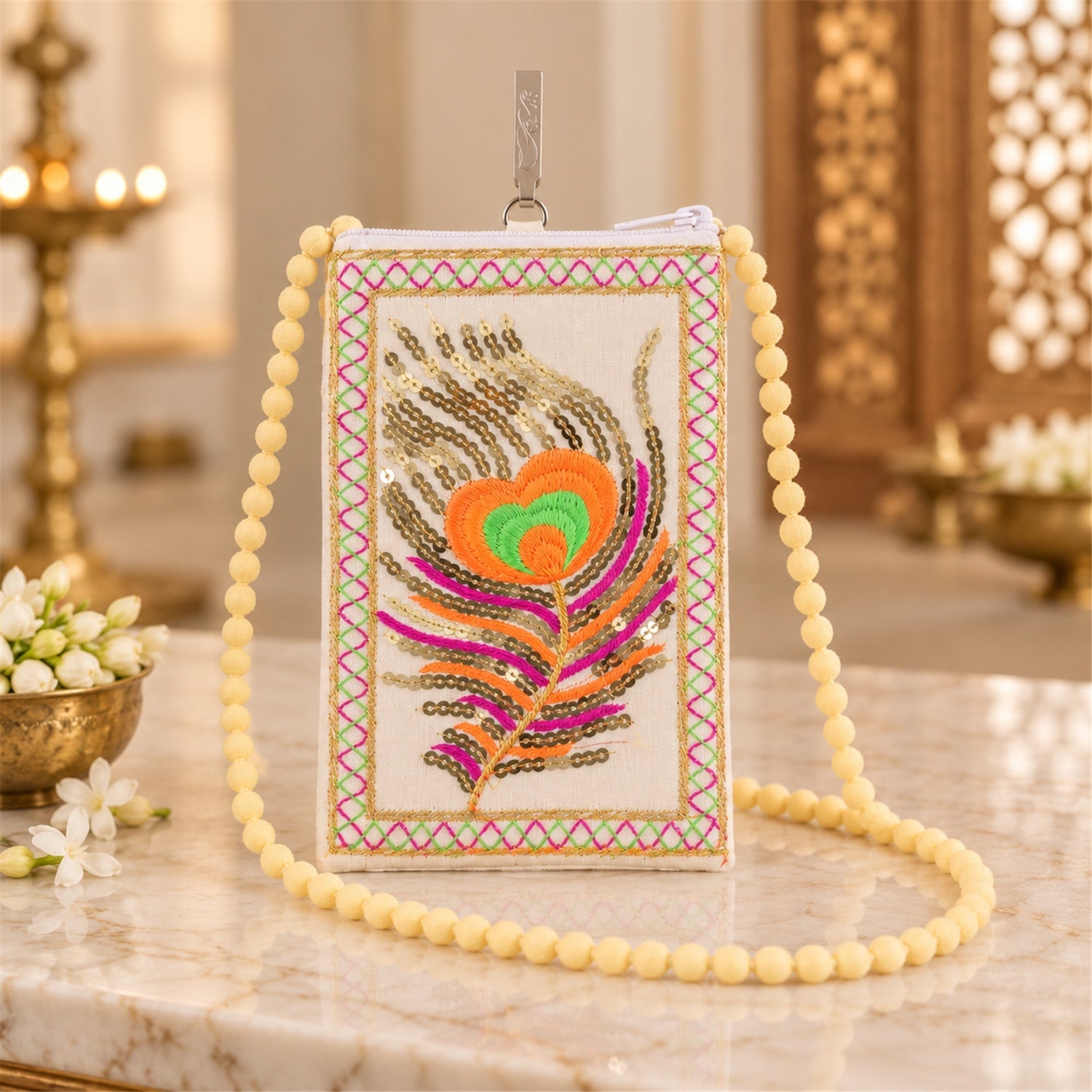 A cream mobile phone pouch with a colourful embroidered peacock feather and a long yellow beaded strap rests on a marble