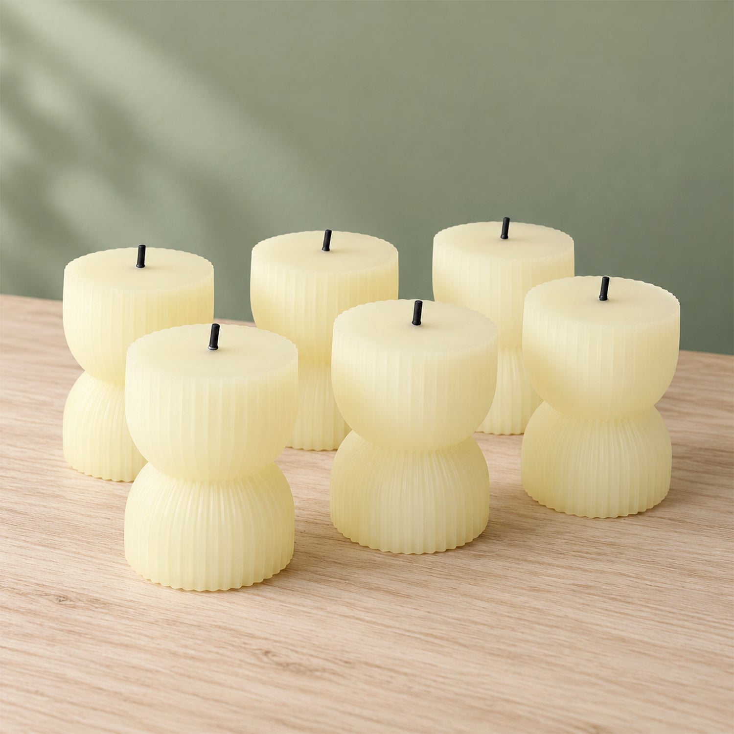 Six unlit cream-colored ribbed LED candles with black wicks arranged on a light wooden surface against a green wall.