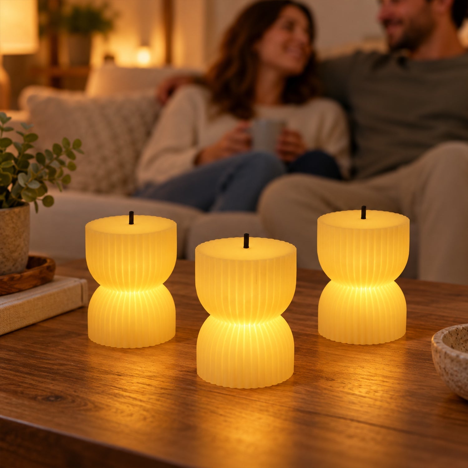 Three glowing ribbed LED candles on a coffee table, with a blurred couple relaxing on a sofa in the background.