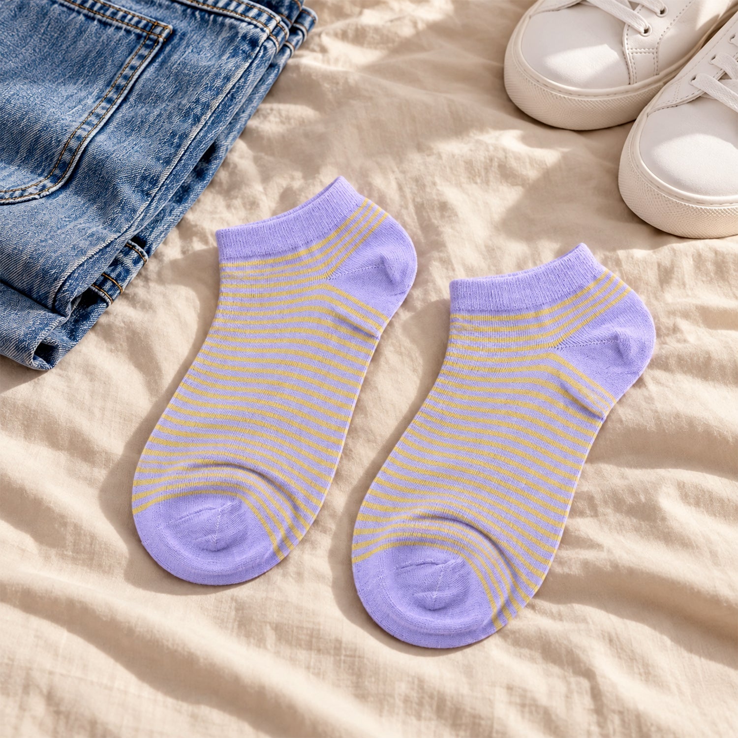 Two light purple and yellow striped ankle socks laid flat on a beige blanket next to blue jeans and white sneakers.