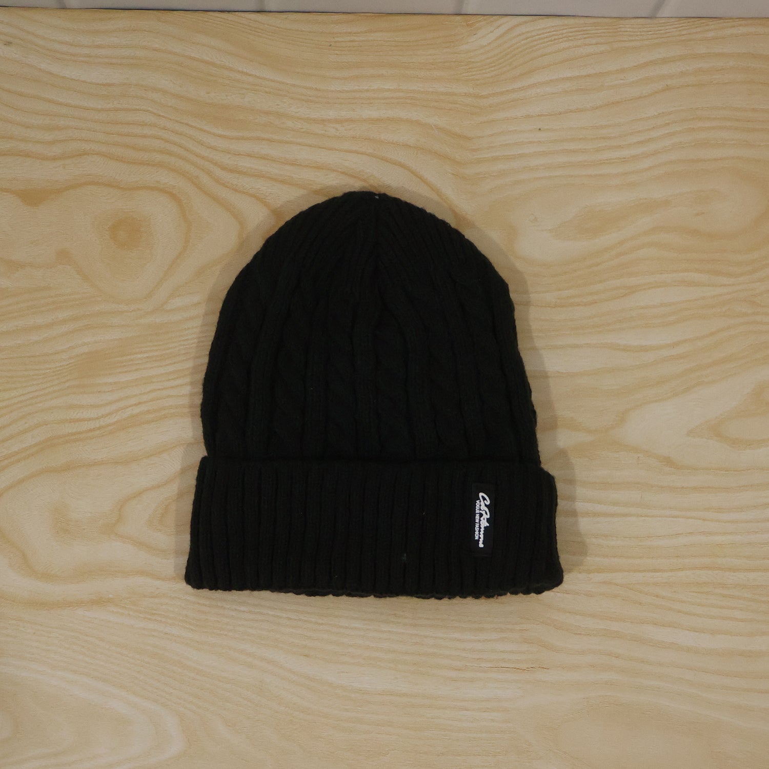 A black cable knit beanie with a ribbed cuff and small white logo, laid flat on a light wooden background.