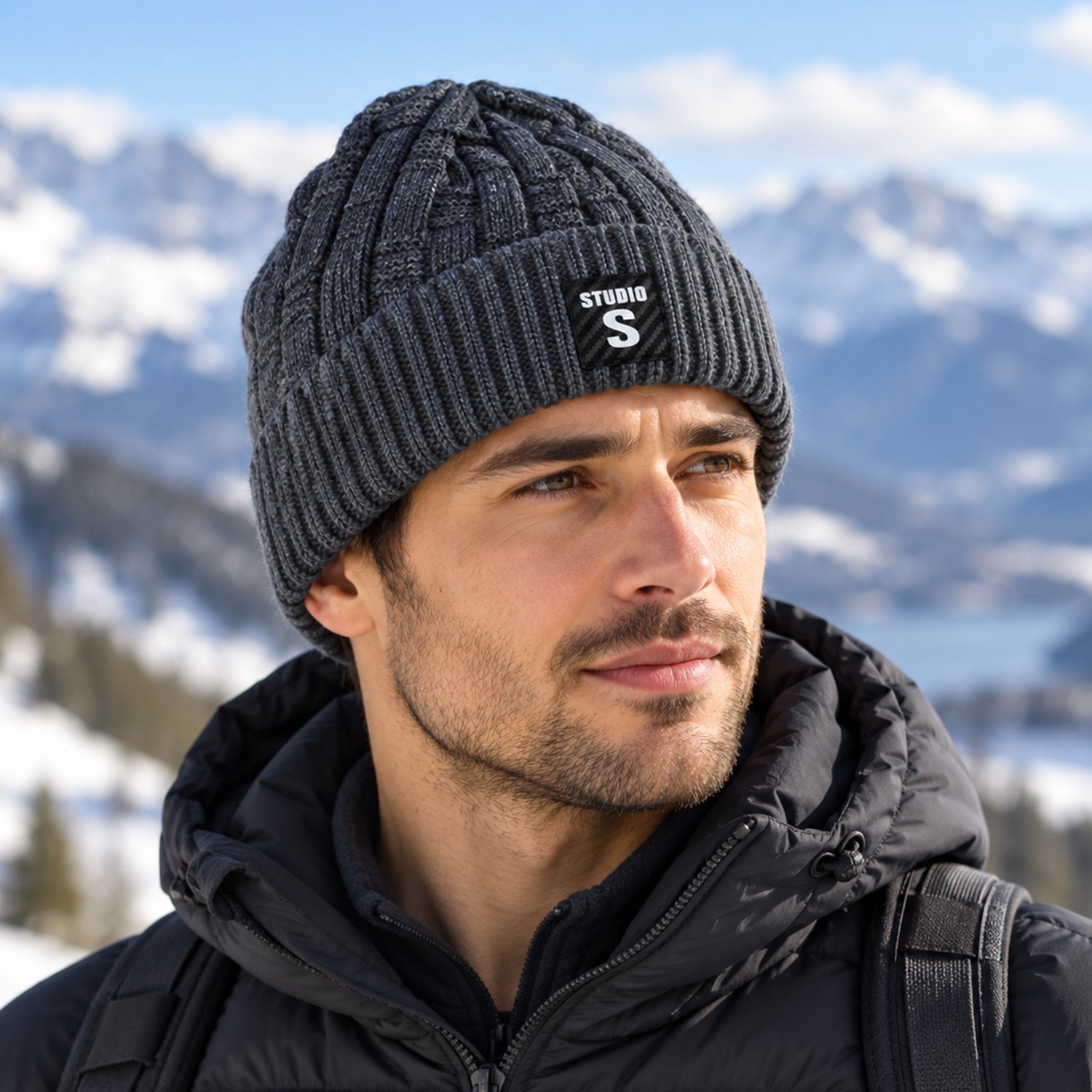 A man wearing a dark grey cable knit beanie with a folded ribbed cuff and a black label, looking to the side in a snowy