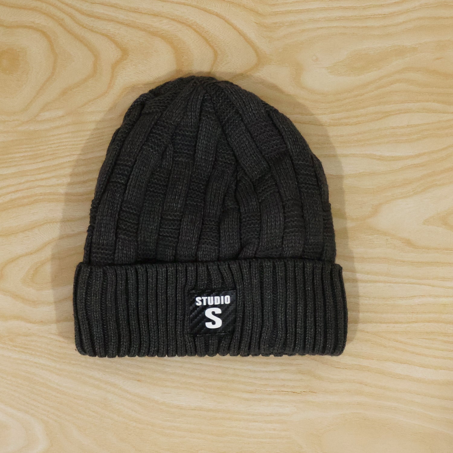 A dark grey cable knit beanie with a ribbed cuff and a black label with "STUDIO S" in white, lying flat on a light wooden