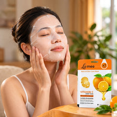 A woman with a white sheet mask on her face gently touches her cheeks, with the product package visible on a table beside