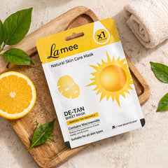 De-tan sheet mask package with a sliced orange and green leaves on a wooden board with a white towel.
