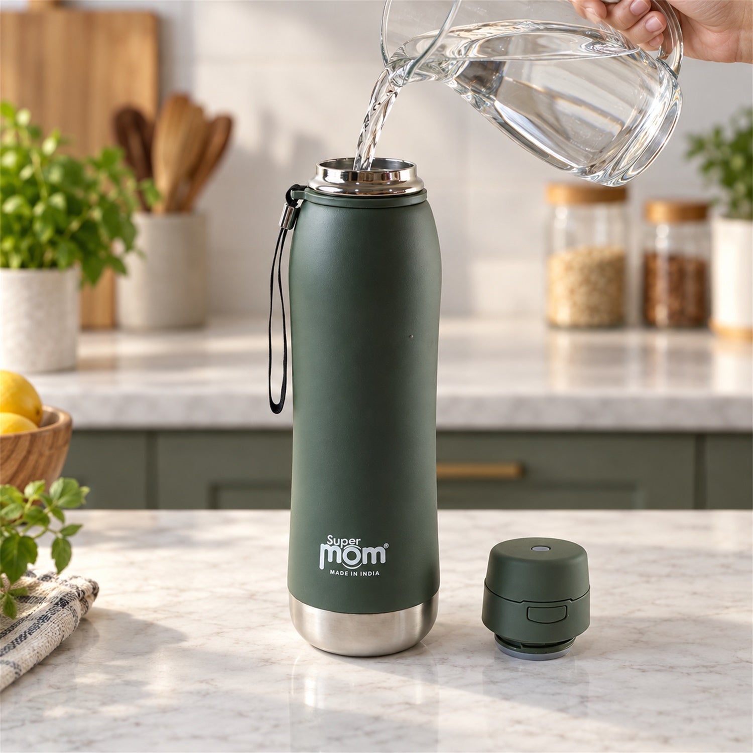 A hand pours water from a clear pitcher into a dark green insulated stainless steel water bottle on a kitchen counter.
