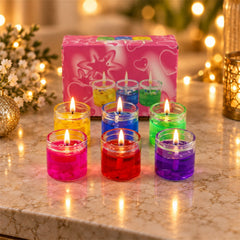 Six lit gel candles in glass jars, in yellow, blue, green, pink, red, and purple, arranged around a pink box.