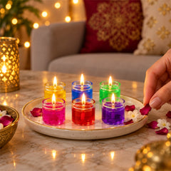 A hand places a pink rose petal on a white plate with six lit gel candles in various colors.