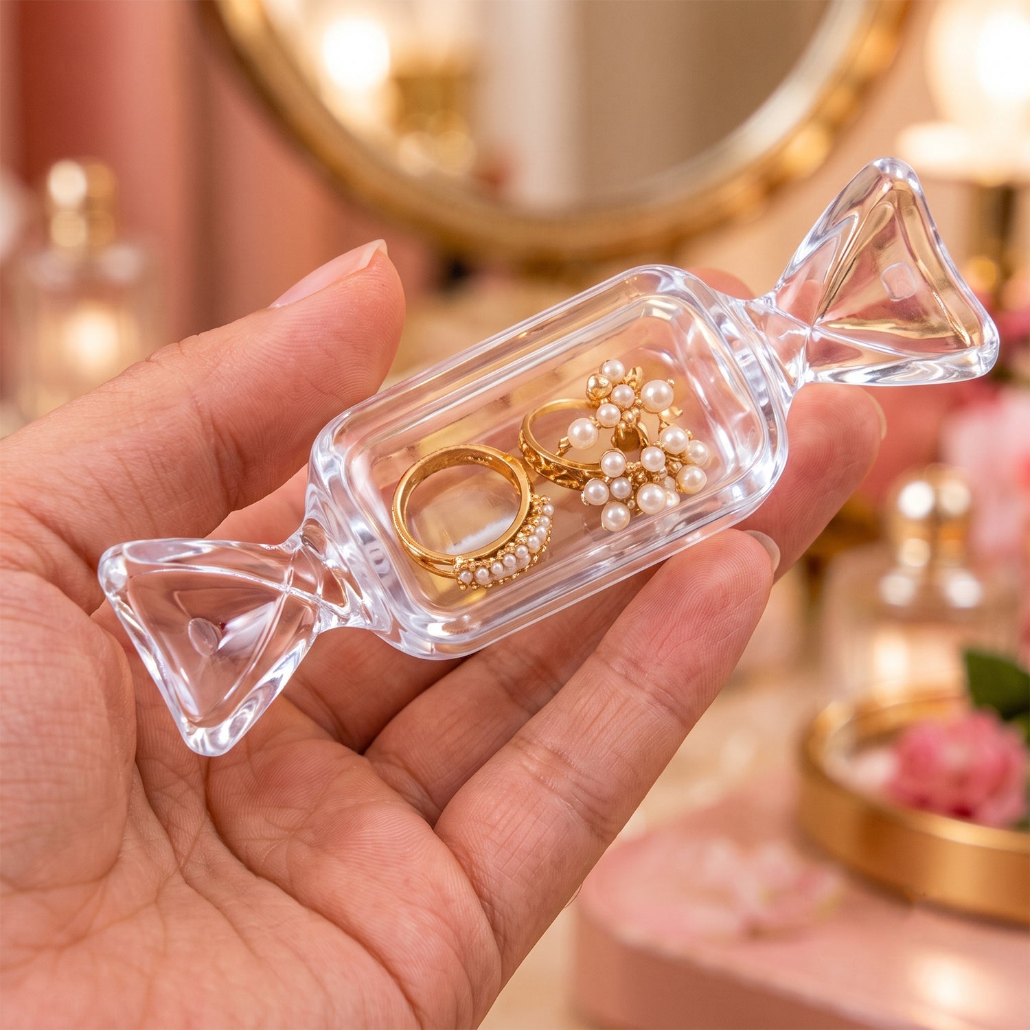 A hand holds the clear candy-shaped box, which contains two gold rings with pearl details, against a blurred vanity