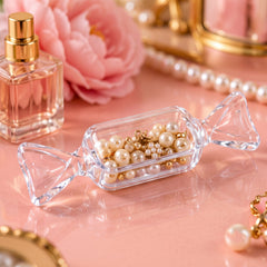A clear, candy-shaped box filled with small, light-colored pearls and gold charms sits on a pink surface next to a perfume
