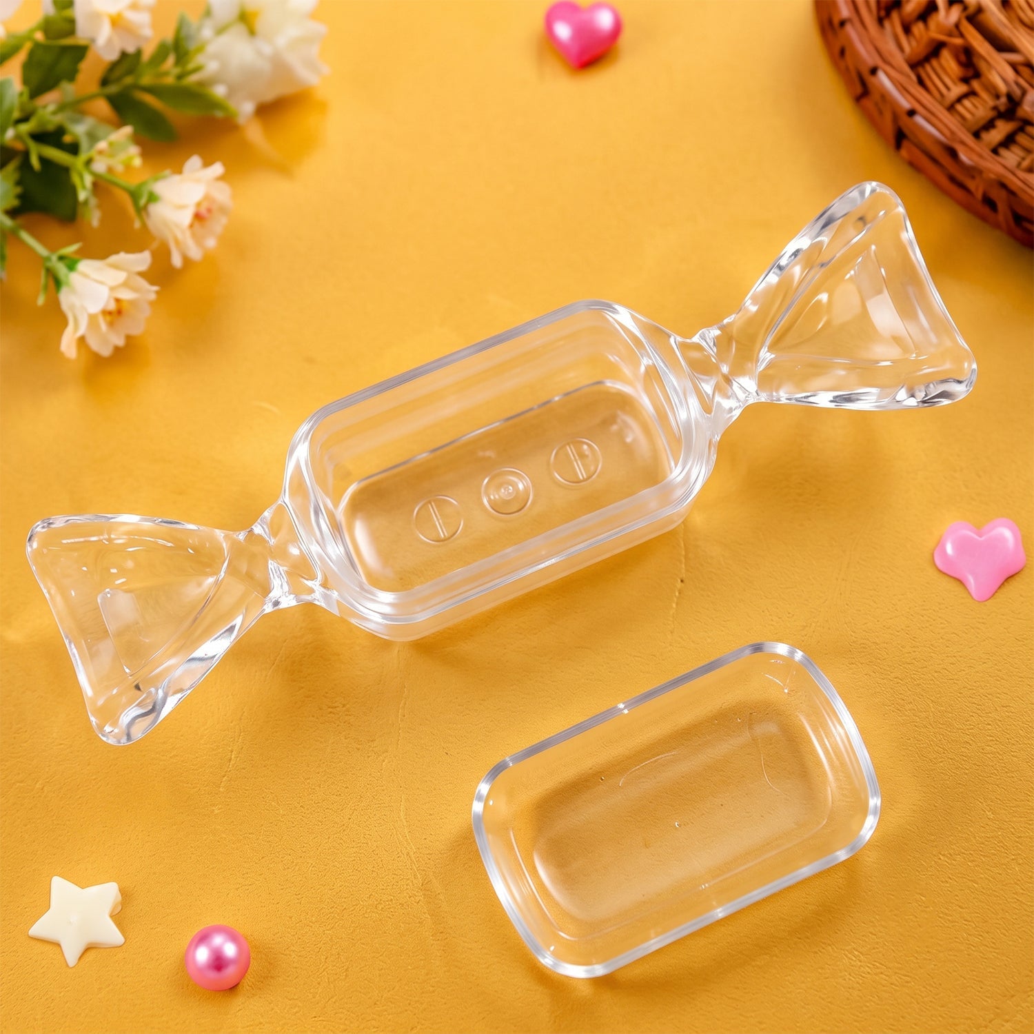The clear candy-shaped box with its lid removed, placed on a yellow surface surrounded by small decorative hearts and