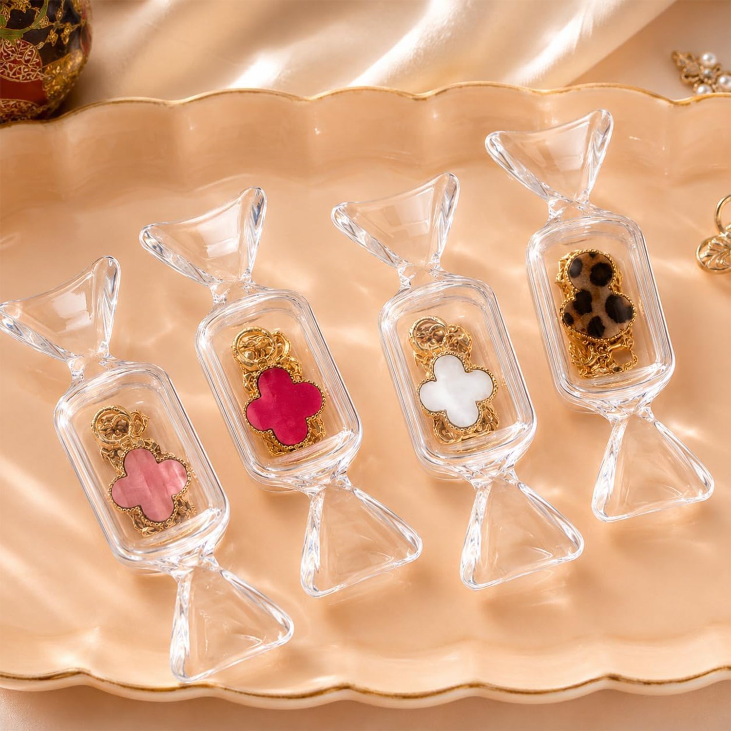 Four clear candy-shaped boxes, each containing a different colored clover-shaped charm, are arranged on a scalloped gold