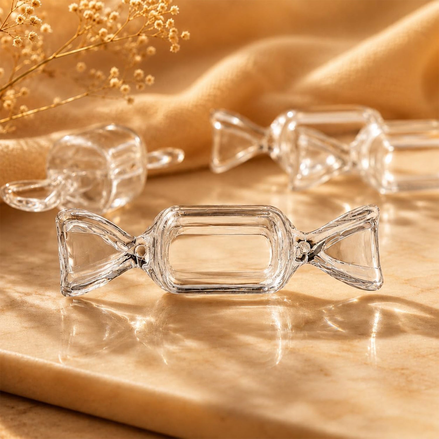 Three clear candy-shaped boxes, two with their lids removed, are arranged on a light brown marble surface with dried foliage.