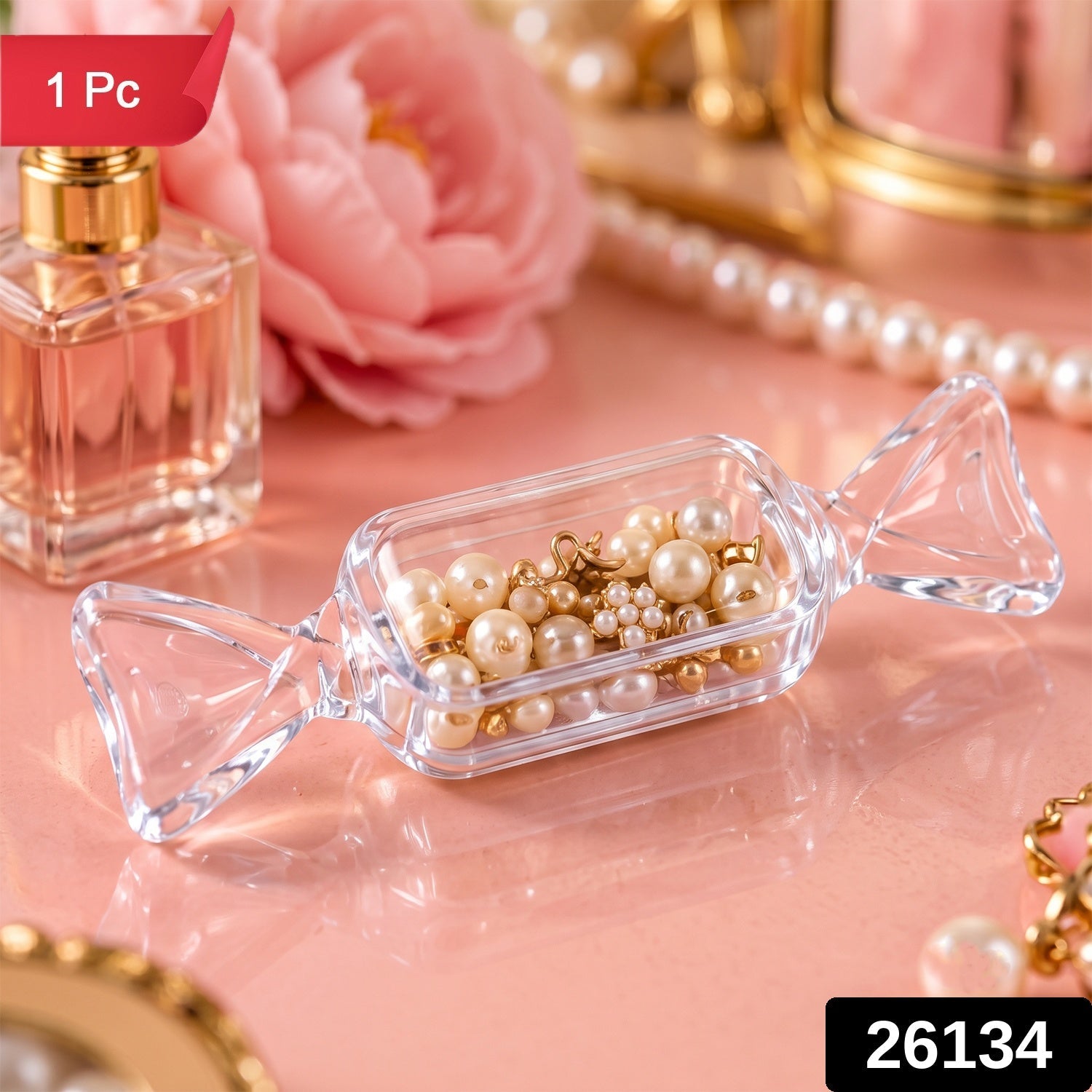 A transparent, candy-shaped container with pearls and gold charms inside, on a pink surface with a perfume bottle and pearl
