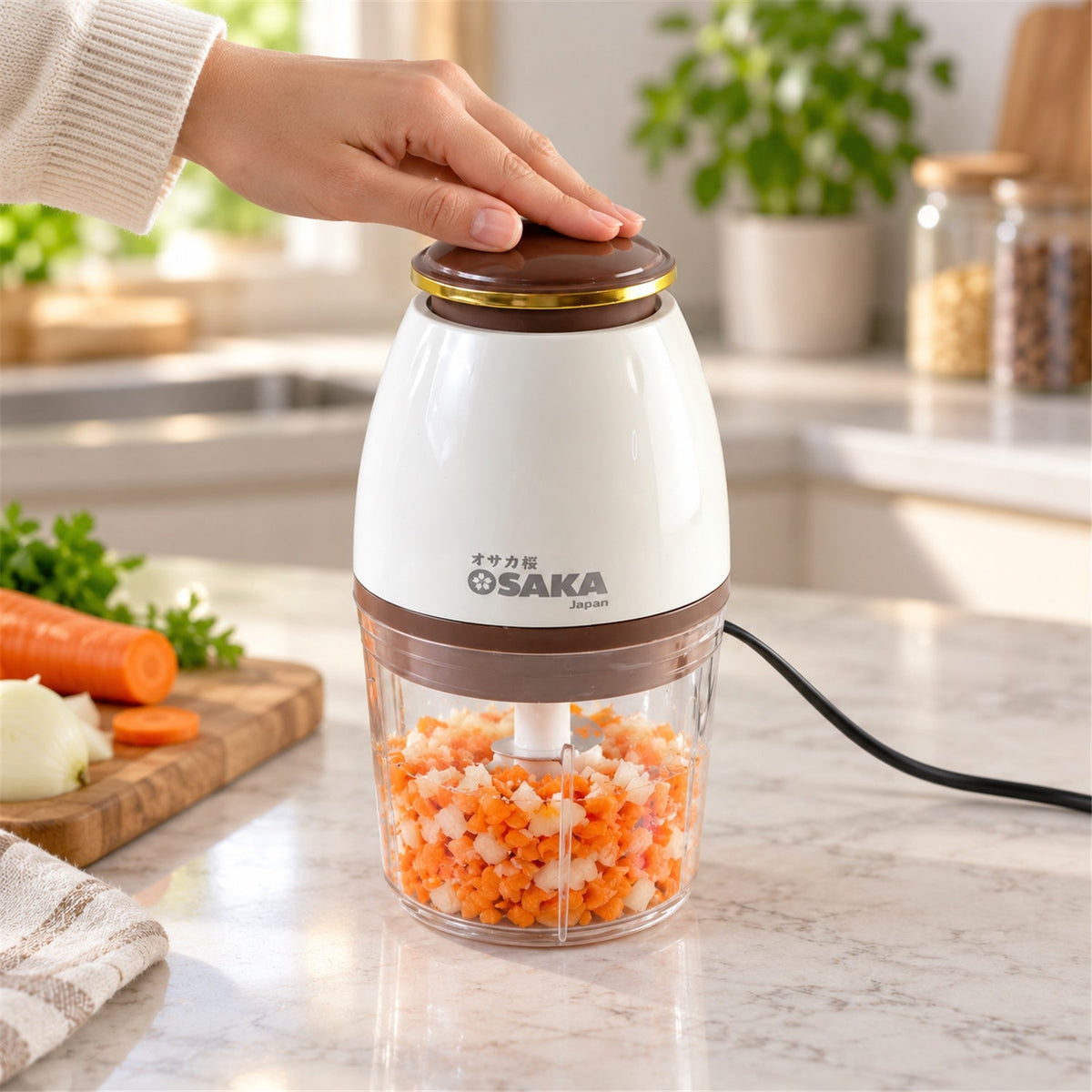 A hand presses the brown top of a white and clear electric chopper filled with diced carrots and onions on a kitchen counter.