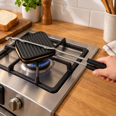 Black stovetop sandwich maker with long metal handles cooking on a gas burner, with a hand holding the handle.