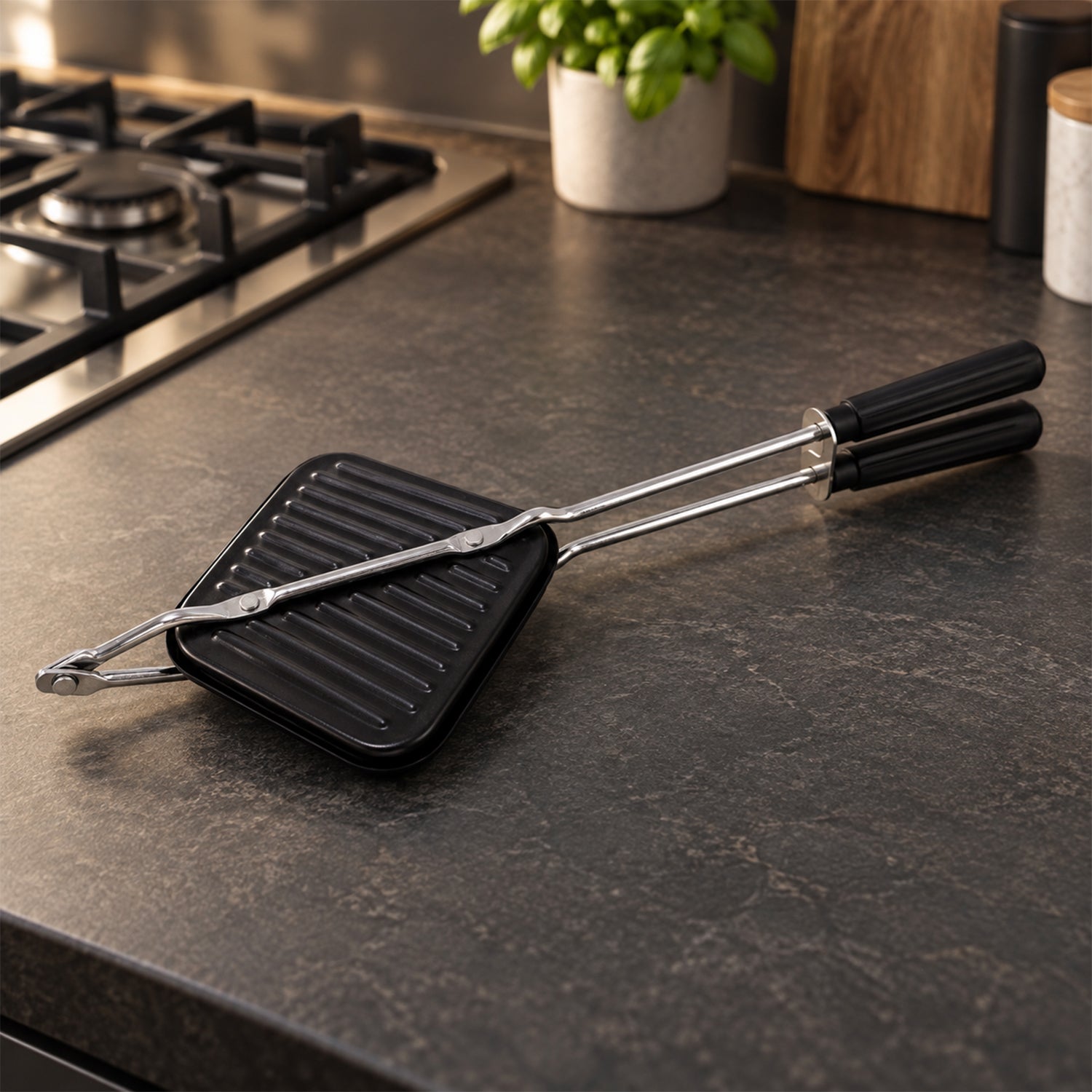 Black stovetop sandwich maker with long silver handles resting on a dark grey speckled kitchen counter next to a gas stove.