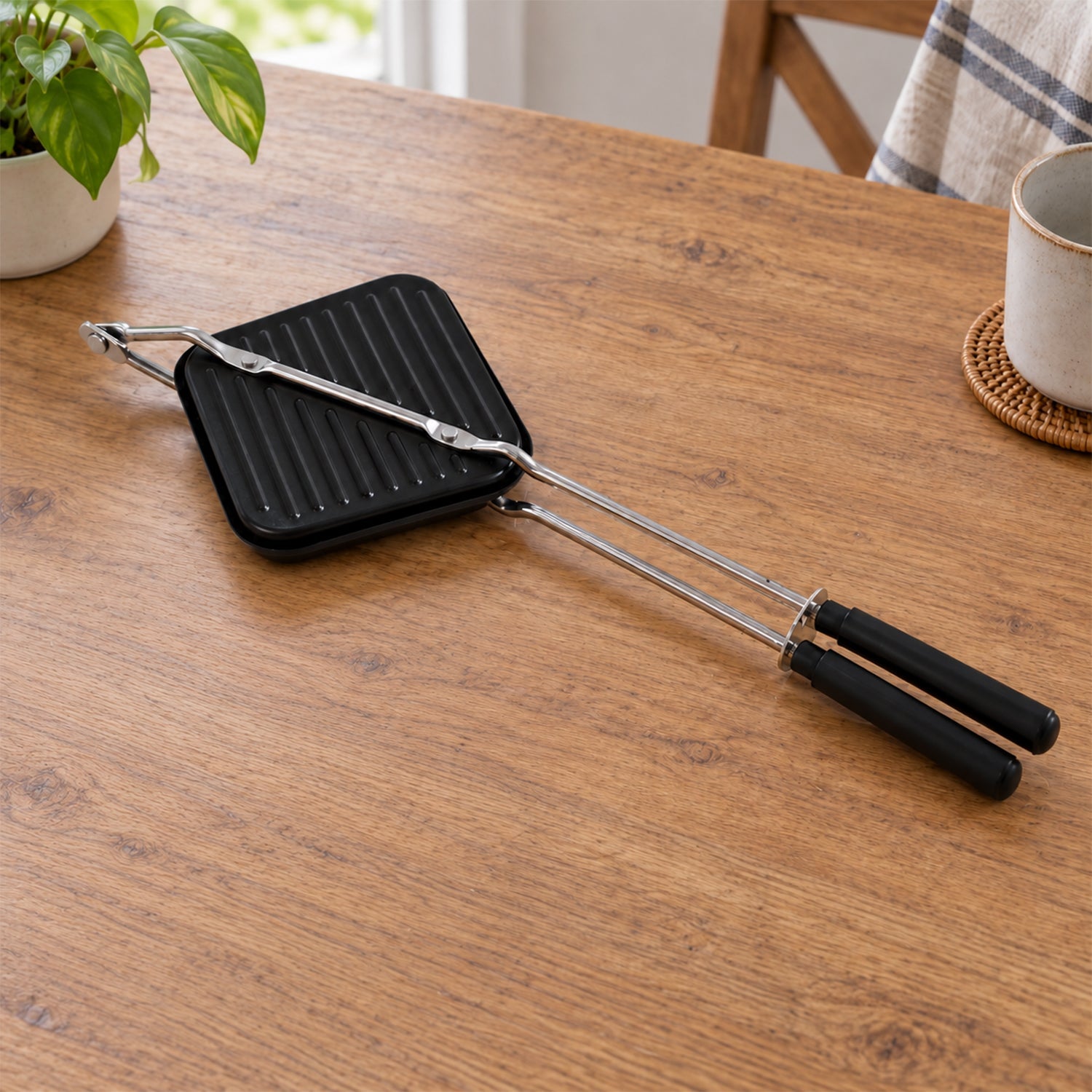 Black stovetop sandwich maker with long metal handles resting on a light brown wooden table, with a plant and mug nearby.