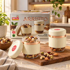 Two clear and cream ribbed containers with rose gold bands, one open, filled with nuts and dried fruit on a wooden board.
