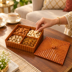A hand reaches into a brown four-compartment dry fruit box, selecting a cashew from the nuts and dried fruits.