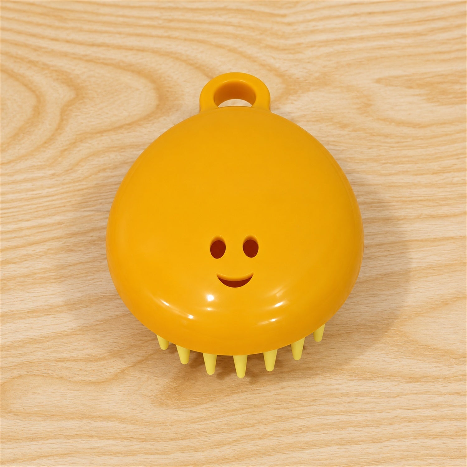 A yellow silicone scalp brush with light green bristles and a smiley face is positioned on a light wooden surface.