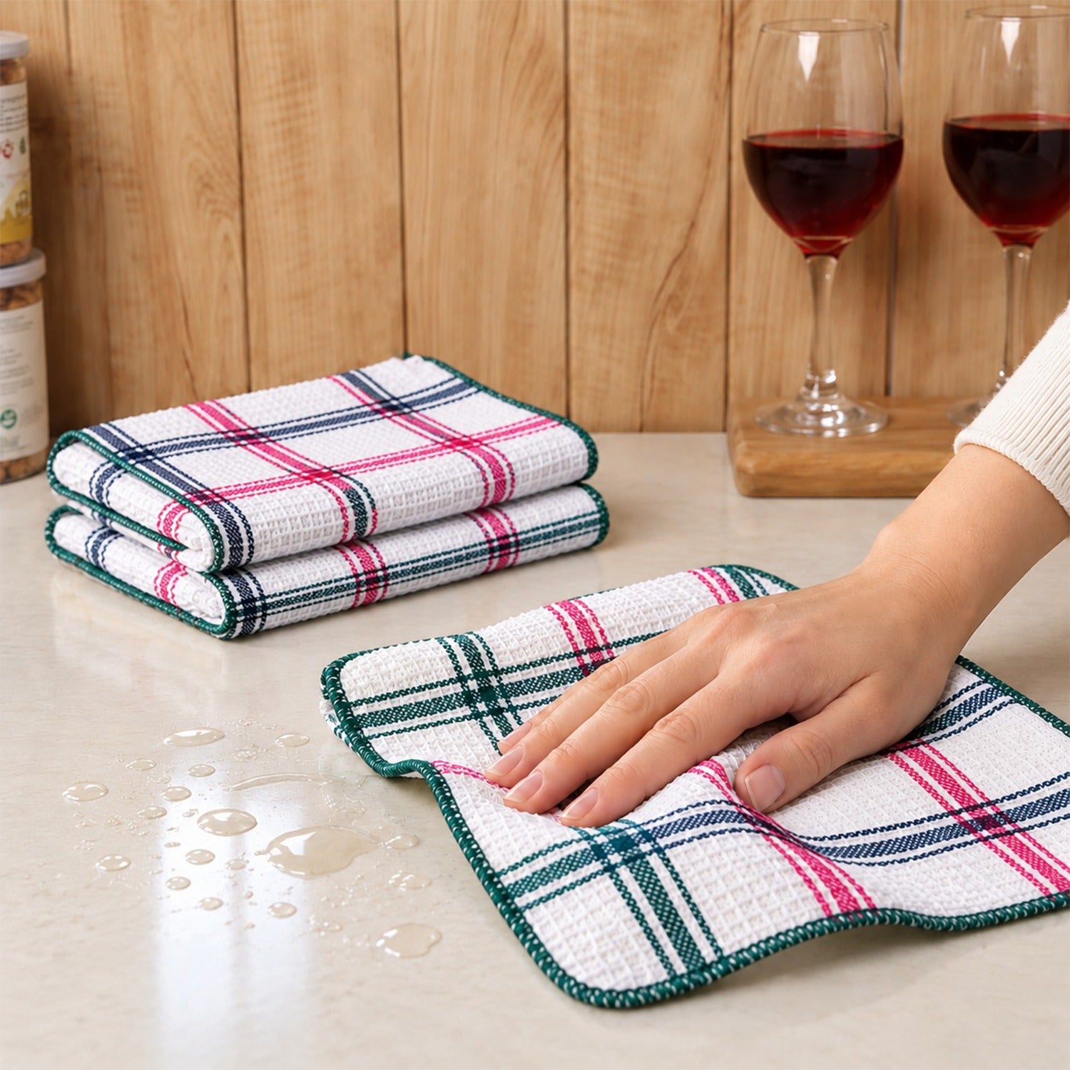 A hand cleans a kitchen counter with a plaid dishcloth, while two wine glasses with red liquid sit nearby.