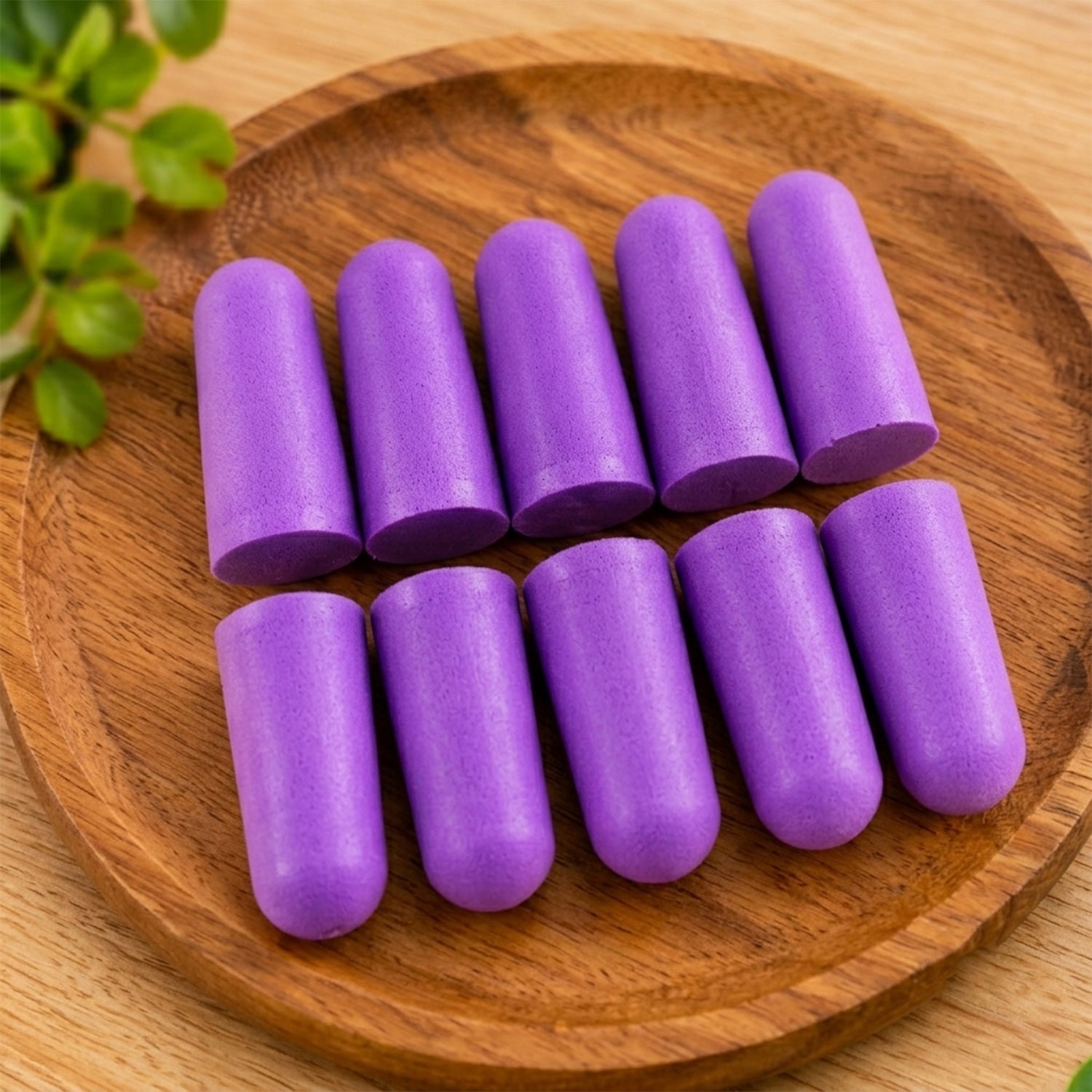 Ten individual purple foam earplugs are arranged in two rows on a round, light brown wooden plate.