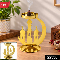 A golden metal incense holder with a crescent moon and mosque design, holding a burning dhoop cone on a wooden table, with a