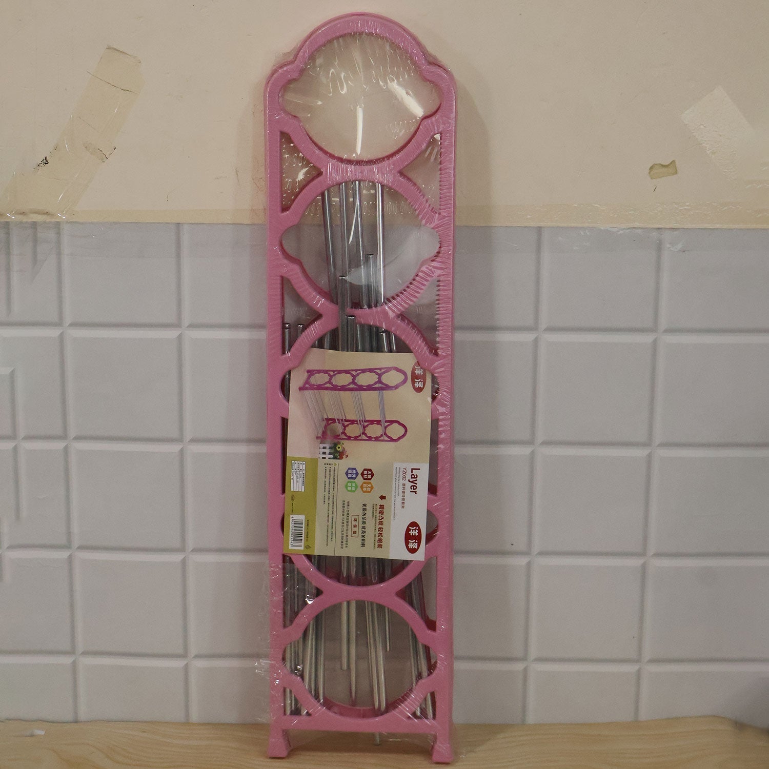 A packaged pink shoe rack, still wrapped in plastic, leaning against a white tiled wall, displaying its compact form before