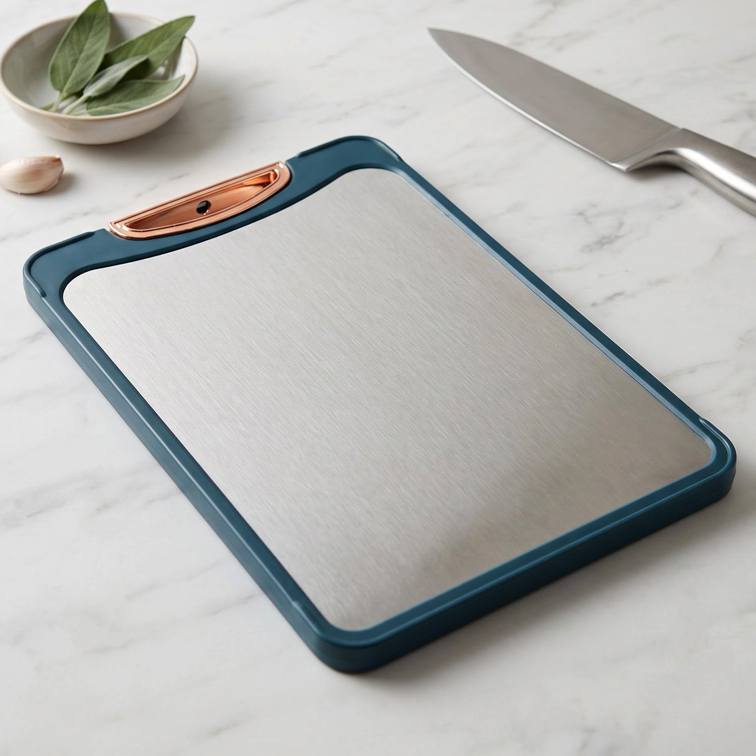 A teal-framed stainless steel cutting board with a copper handle sits on a white marble counter next to a knife.