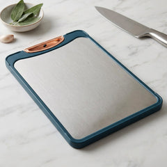 A teal-framed stainless steel cutting board with a copper handle sits on a white marble counter next to a knife.