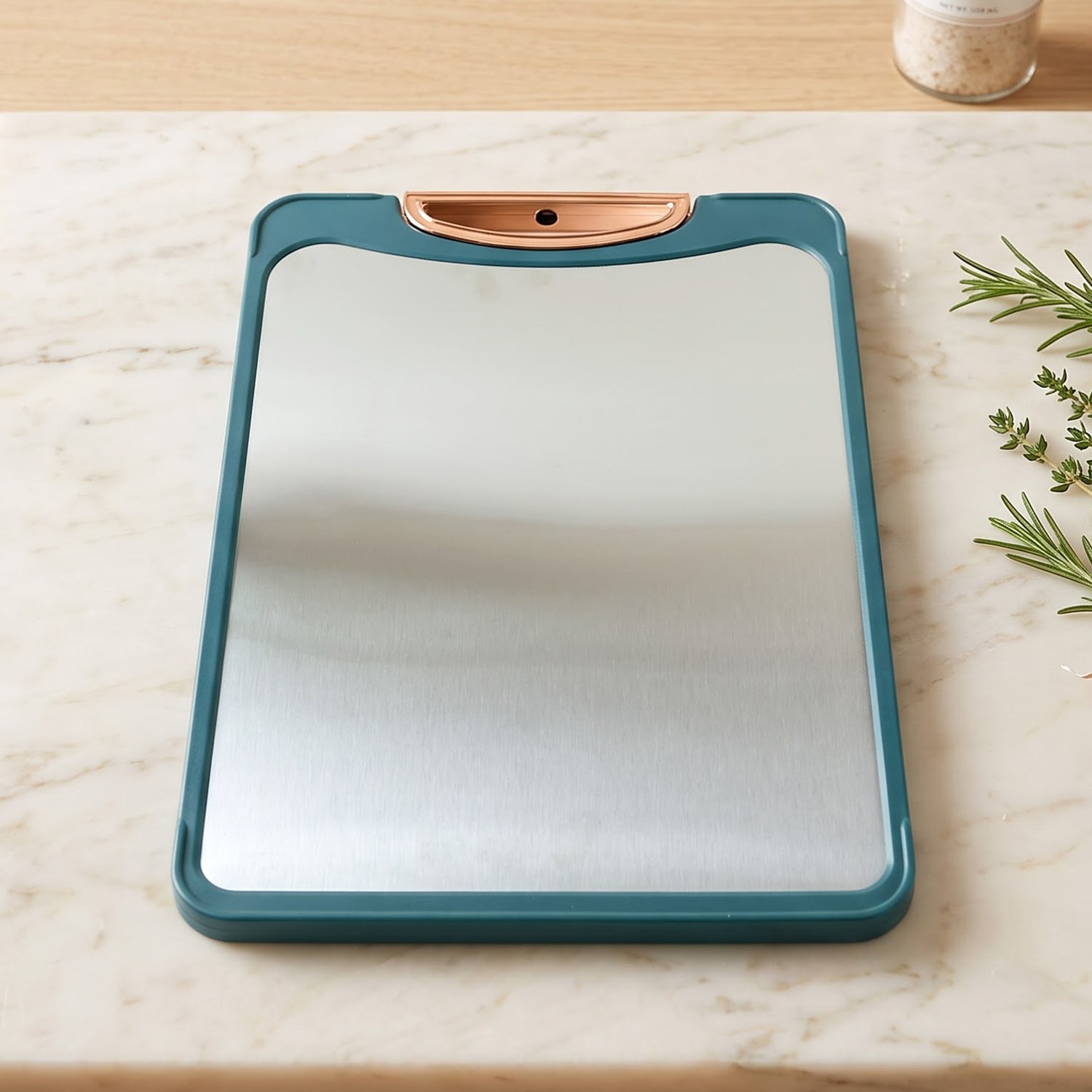 A teal and stainless steel cutting board with a unique copper handle rests on a light marble surface.