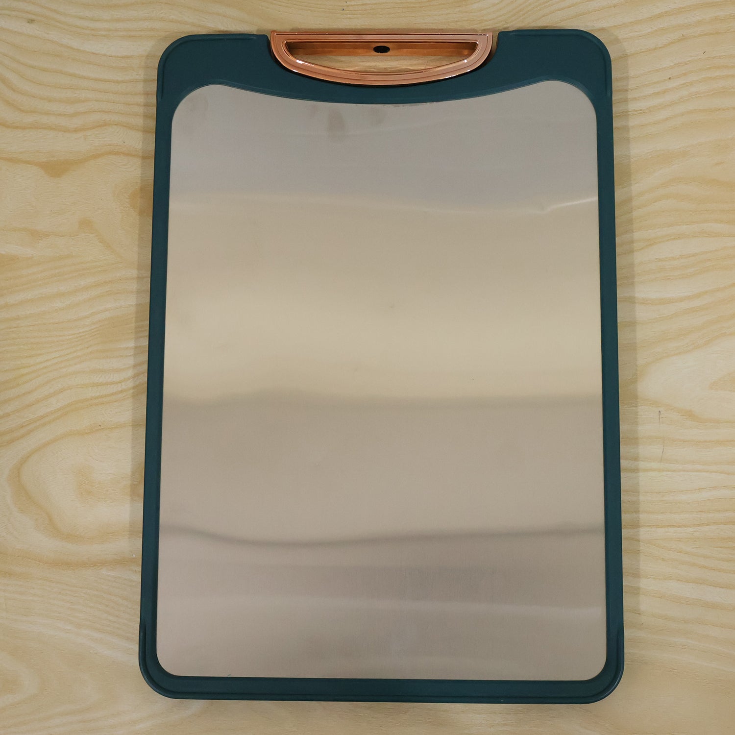 A teal-edged stainless steel cutting board with a curved copper handle lies flat on a light wood surface.