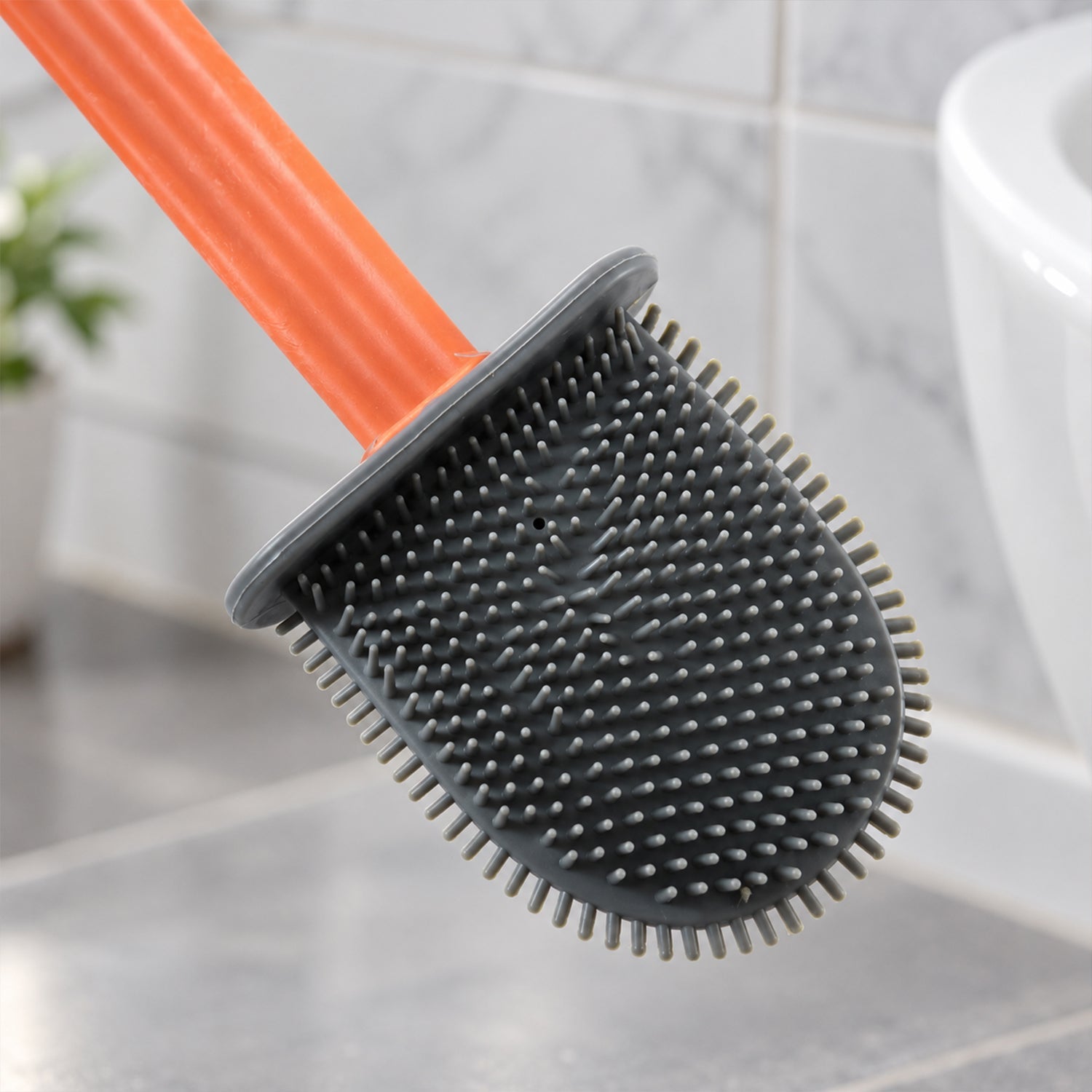 A close-up view of the grey silicone brush head with numerous flexible bristles and an orange handle, near a white toilet.