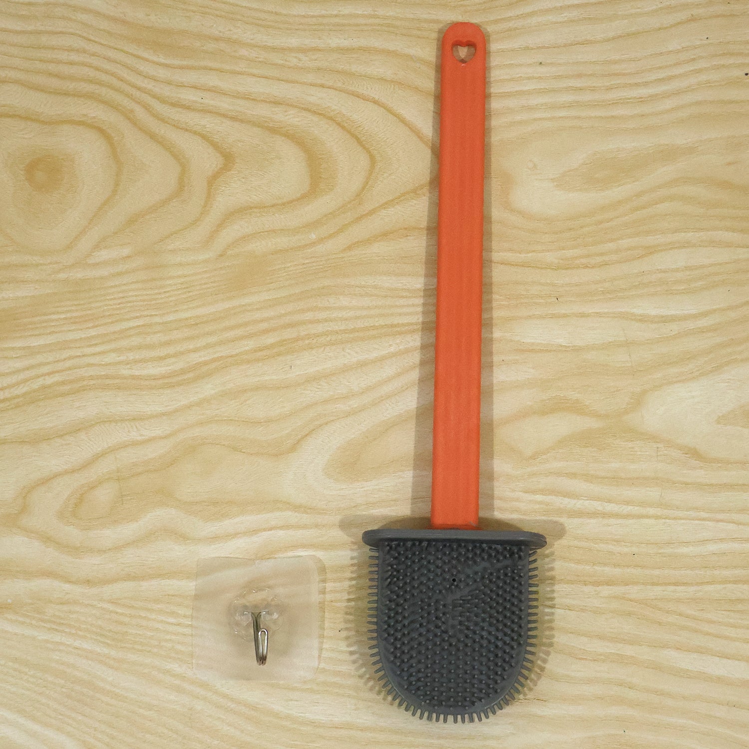 An orange-handled, grey silicone toilet brush and a clear adhesive wall hook lie flat on a light wooden surface.