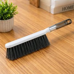 A white and gray cleaning brush with dark bristles rests on a light wooden surface next to a small potted plant.