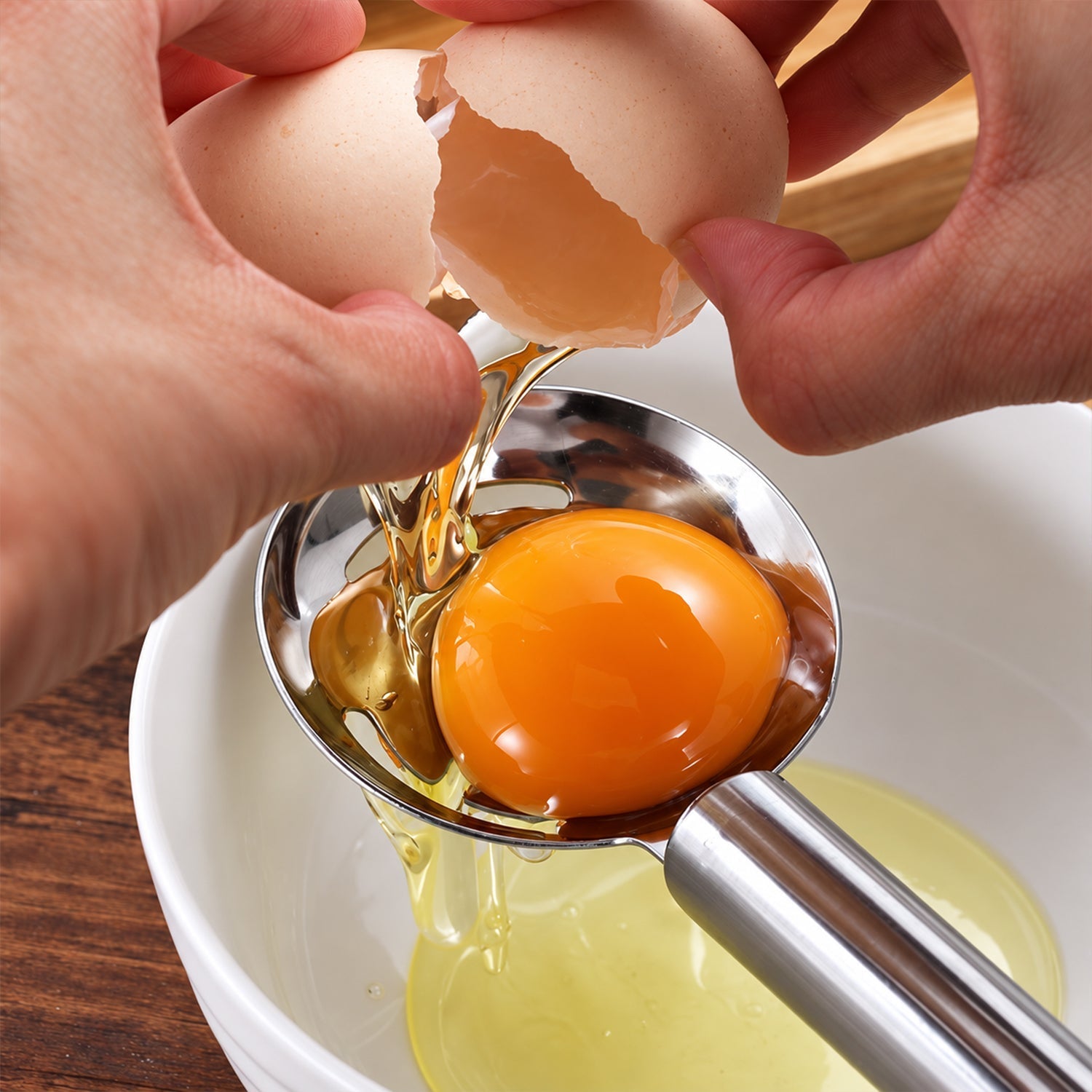 Hands crack an egg directly into the stainless steel separator, allowing the white to drip into a white bowl.