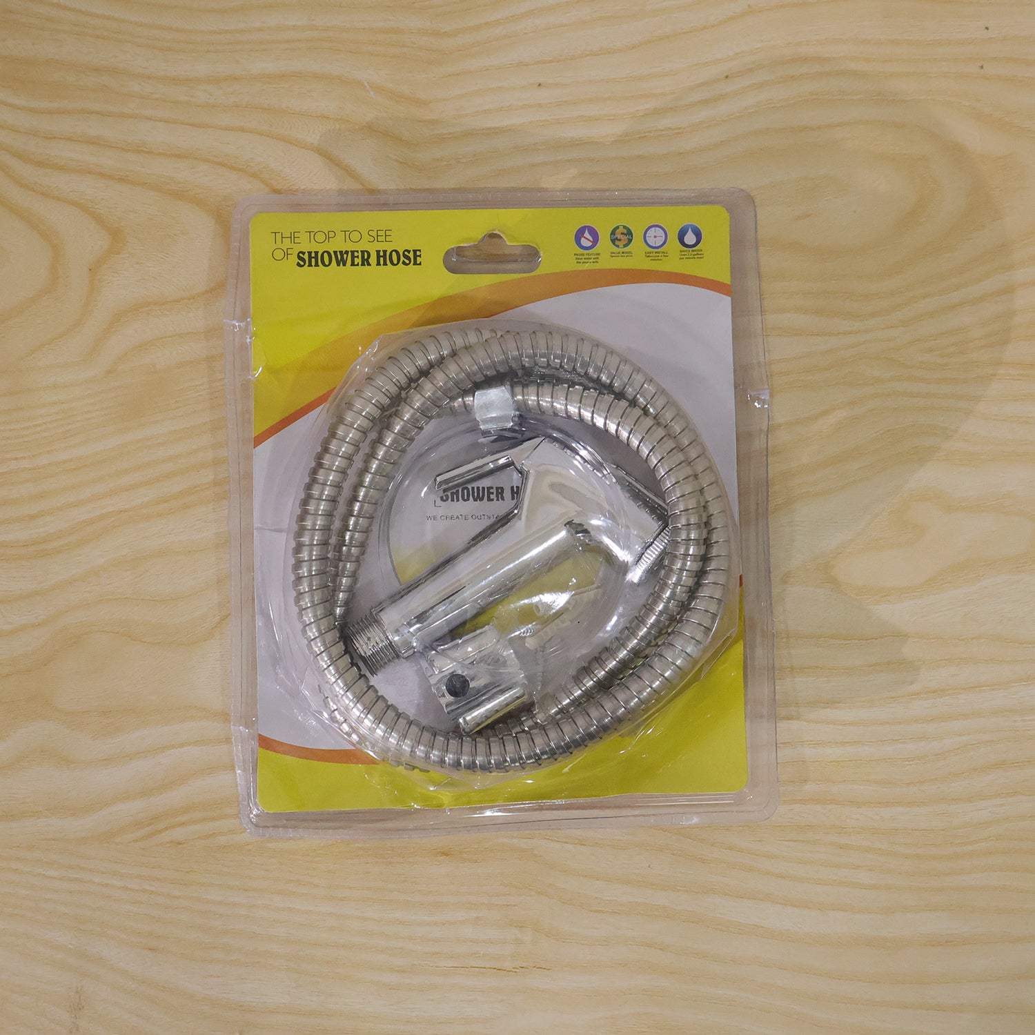 A packaged chrome bidet sprayer kit with a coiled hose is sealed in clear plastic on a light wooden surface.