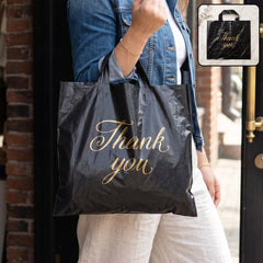 A person in a denim jacket holds a black plastic 'Thank You' bag with gold script, outdoors.