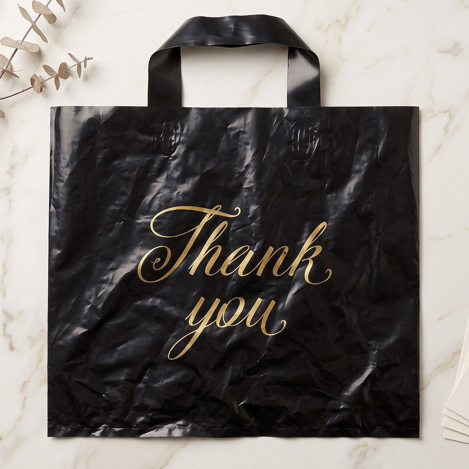A black plastic 'Thank You' bag with gold script lies flat on a white marble surface with eucalyptus leaves.