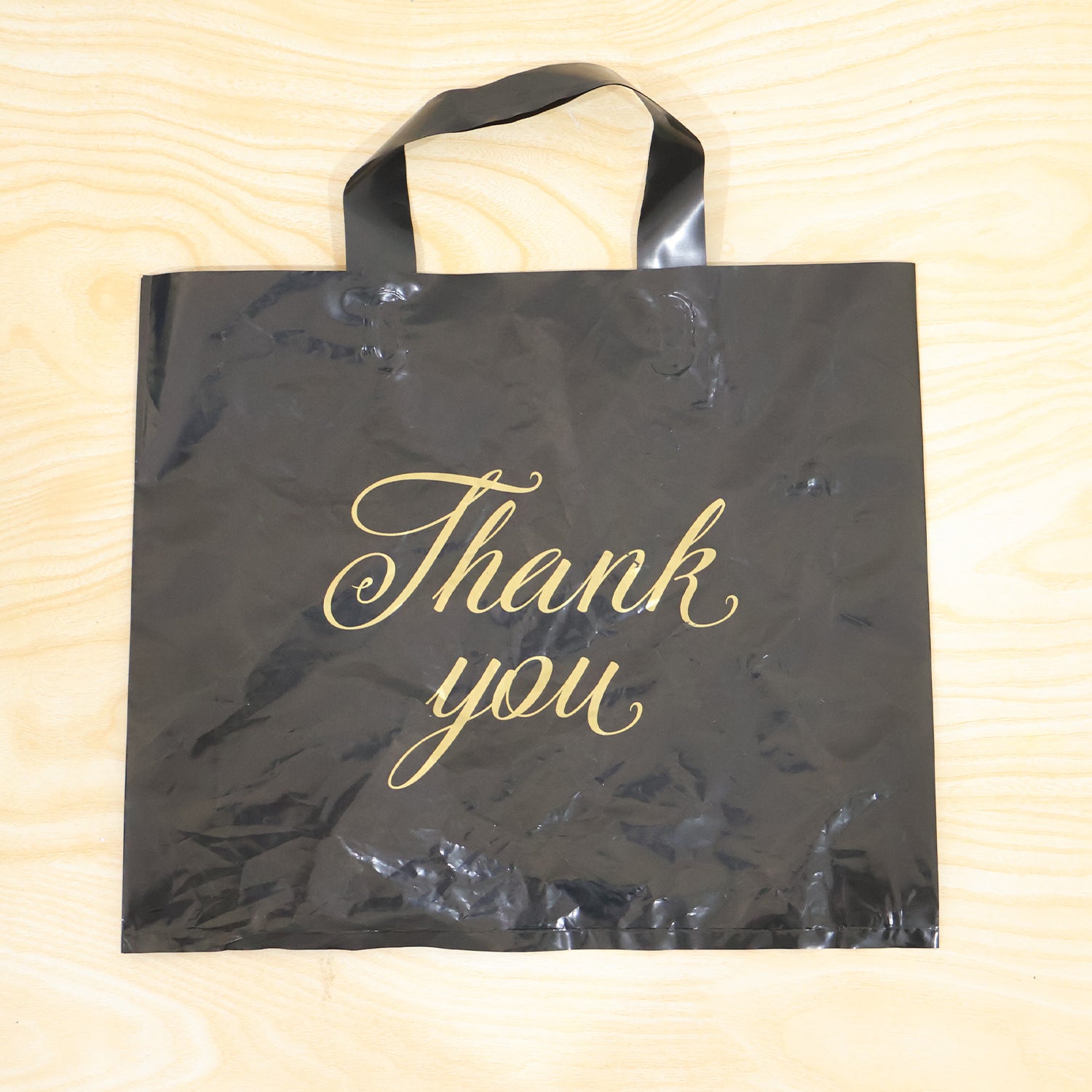 A black plastic 'Thank You' bag with gold script is displayed flat on a light-colored wooden background.