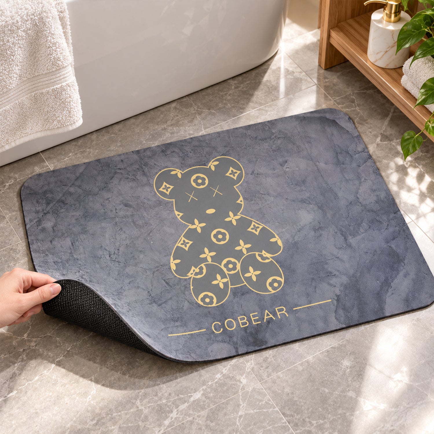 A hand lifts the corner of the grey bath mat, revealing its textured anti-slip backing on a tiled floor.