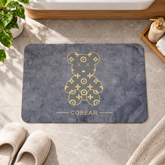 A grey rectangular bath mat with a golden bear design lies on a tiled floor next to beige slippers.