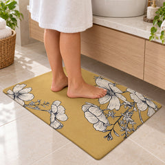 A person in a white robe stands on a mustard yellow mat adorned with white floral illustrations in a bathroom.