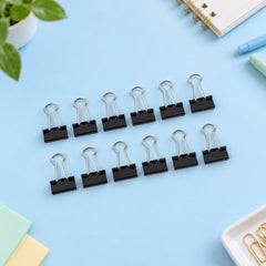 Twelve black binder clips with silver handles are neatly arranged in rows on a light blue surface with office supplies.